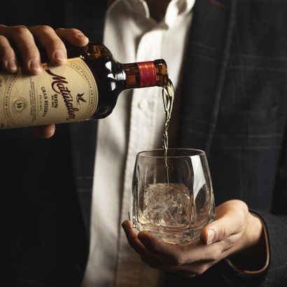 Ein eleganter Mann serviert Rum in ein Glas, das mit Eis gefüllt ist, und zeigt dabei eine entspannte, genussvolle Haltung.
