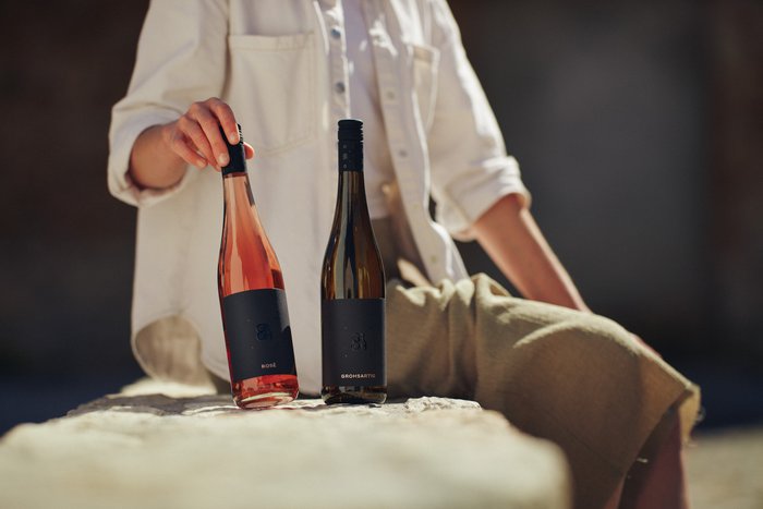 Eine Person hält eine Flasche Roséwein und eine Flasche Weißwein, während sie entspannt auf einem Stein sitzt.