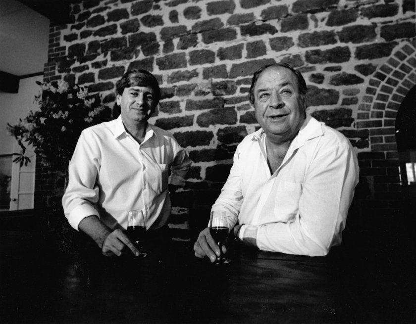 In einer einladenden Atmosphäre mit einer Steinwand lächeln zwei Männer, während sie Gläser in der Hand halten und sich unterhalten.