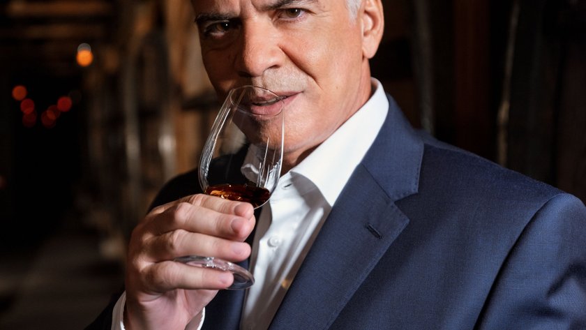 In einem gedämpften Lichtumfeld steht ein Mann in einem eleganten Anzug und nippt an seinem Whisky, während er aufmerksam schaut.