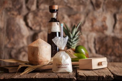 Der rustikale Tisch zeigt eine Kokosnuss, eine Flasche und eine Cocktailtüte, umgeben von frischen Ananas und Limetten.