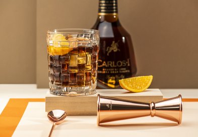 Im Hintergrund ist eine Flasche mit der Aufschrift 'CARLOS I' zu sehen, während ein Messbecher aus Kupfer im Vordergrund liegt.