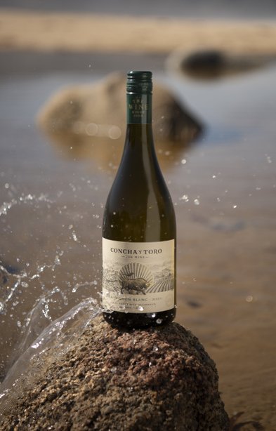 Die Flasche Wein von Concha y Toro ist auf einem Felsen platziert, umgeben von spritzendem Wasser und einem sanften Strandhintergrund.