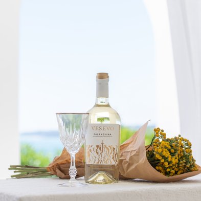 Ein eleganter Tisch mit einer Flasche Vesevo Falanghina, einem Kristallglas und einem Strauß gelber Blumen in braunem Papier.