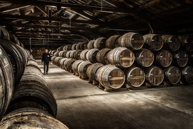 Ein Mann geht durch einen dunklen Lagerraum, umgeben von Reihen alter Holzfässer, die sorgfältig aufgestellt sind.