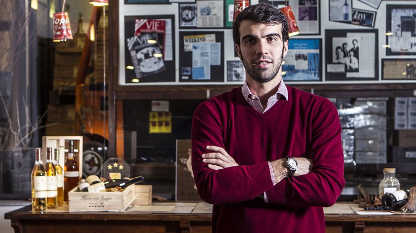 Der Mann in einem roten Pullover lächelt leicht, während er vor einem Tisch mit Weinflaschen und Holzboxen posiert.