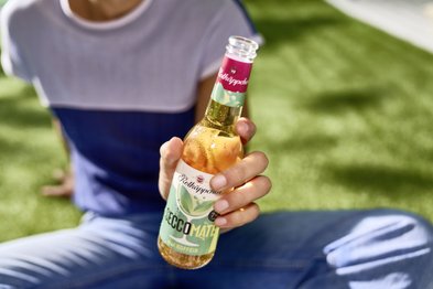 In der Hand einer Person glänzt eine Flasche Secco Mate, umgeben von einer sonnigen, grünen Umgebung.