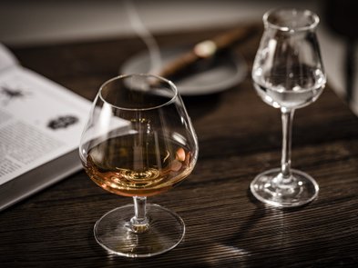 Ein elegantes Glas mit goldfarbenem Getränk steht auf einem Holztisch, daneben ein weiteres Glas mit klarem Inhalt.