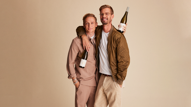 Zwei Männer stehen eng umarmt vor einem neutralen Hintergrund und halten Flaschen Wein in der Hand, lächelnd und glücklich.