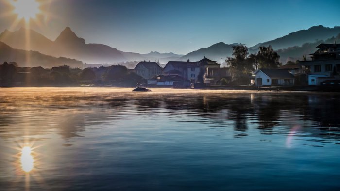 Die Sonne strahlt über einen ruhigen See, während Nebel sanft über das Wasser schwebt und die umliegenden Berge umhüllt.