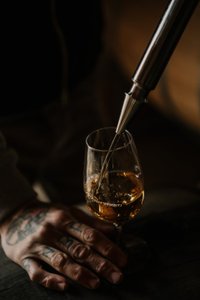 In einem gedämpften Licht wird ein Glas mit einem bernsteinfarbenen Getränk von einer Hand mit Tattoos gehalten.