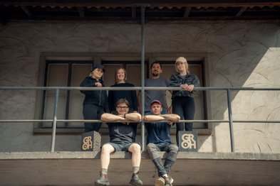 Sechs Personen stehen und sitzen auf einem Balkon, lächeln und posieren entspannt in der Sonne.