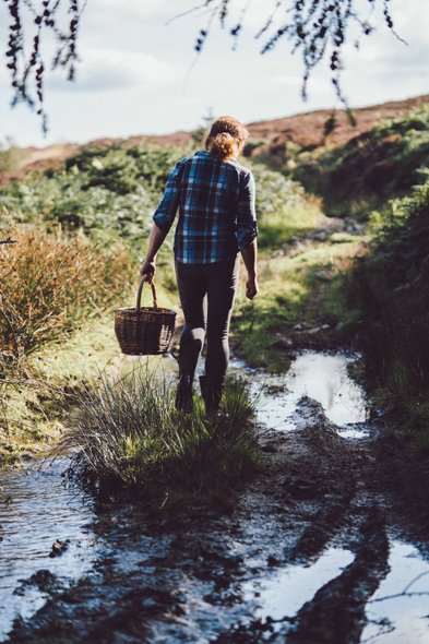 Eine Person mit einem Korb geht einen schlammigen Weg entlang, umgeben von üppigem Grün und sanften Hügeln im Hintergrund.