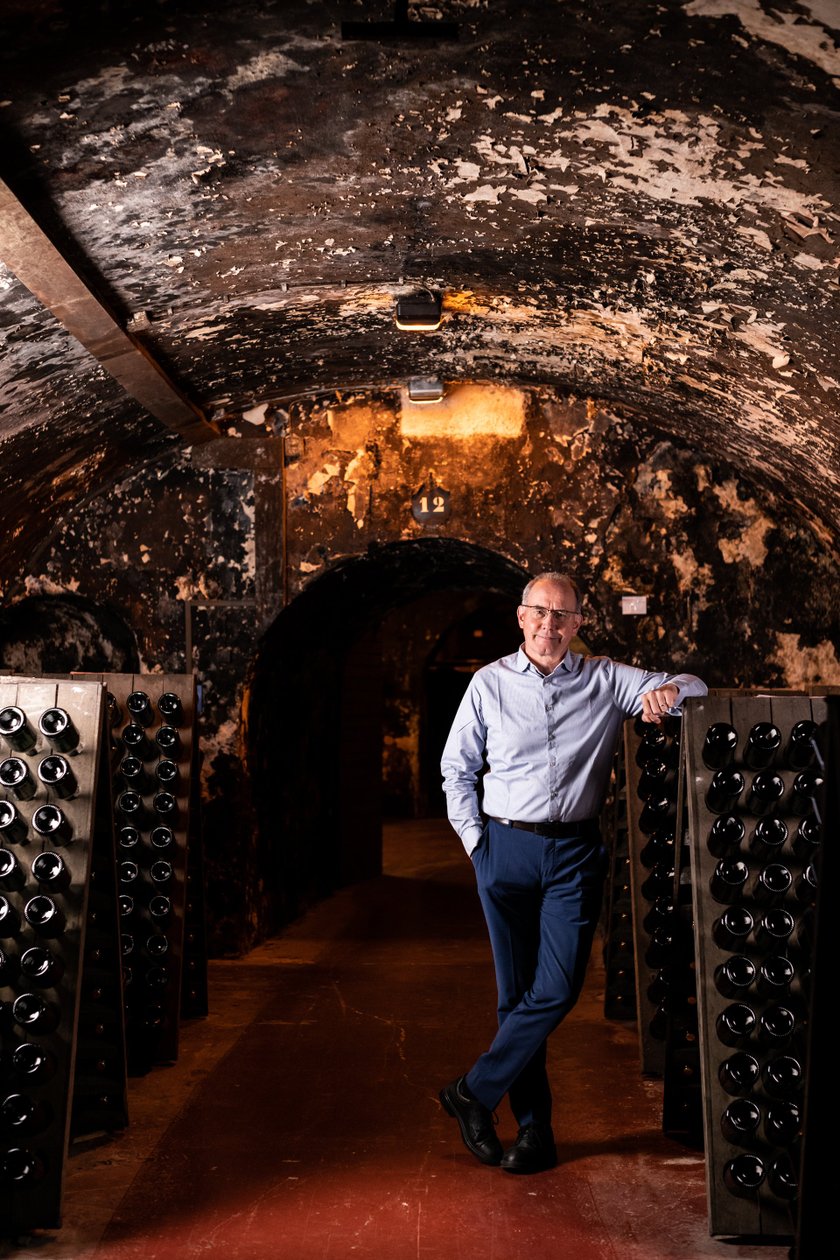 Ein Mann steht entspannt in einem Weinkeller, umgeben von Regalen mit dunklen Weinflaschen und einer rustikalen, abgenutzten Wand.