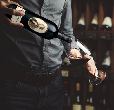 Ein Mann in einem grauen Hemd gießt roten Wein aus einer Flasche in ein Glas, während er eine entspannte Haltung einnimmt.