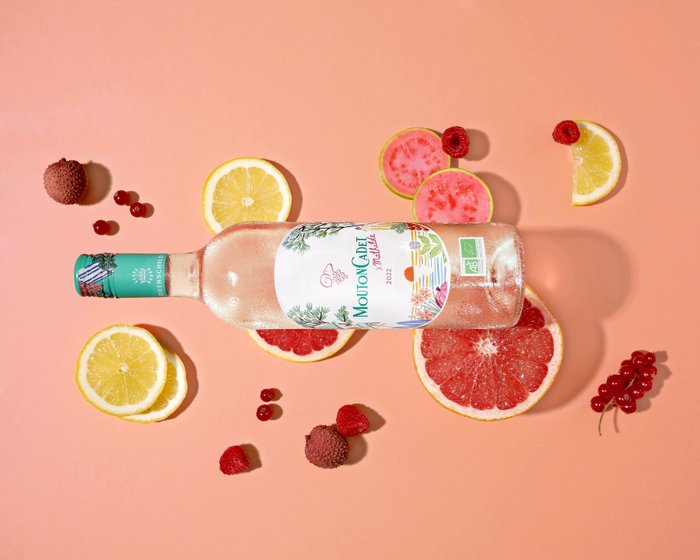 Eine Flasche Mouton Cadet Rosé liegt auf einem rosa Hintergrund, umgeben von frischen Früchten wie Grapefruit, Zitrone und Himbeeren.