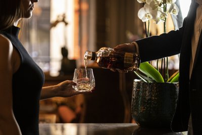 In einem luxuriösen Setting wird einer Frau von einem Mann ein Getränk in ein Glas eingeschenkt, umgeben von Pflanzen.