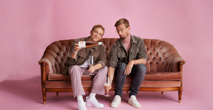 In einem rosa Hintergrund sitzen zwei Männer auf einem Sofa, einer lächelt beim Einschenken von Wein, der andere zeigt eine nachdenkliche Miene.