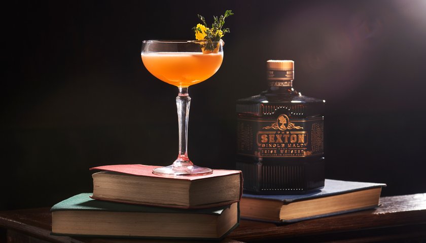 Ein elegantes Cocktailglas ist mit einer orangen Flüssigkeit befüllt und steht auf einem Bücherstapel neben einer Flasche "The Sexton" vor einem dunklen Hintergrund.