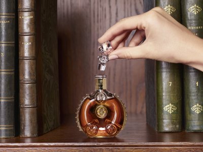 Eine Hand hebt den kunstvoll gestalteten Korken einer eleganten Flasche, die auf einem Holzregal zwischen alten Büchern steht.