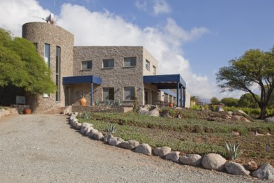 Ein modernes, steinernes Gebäude mit blauen Akzenten steht vor einem klaren Himmel, umgeben von üppigem Grün und Felsen.