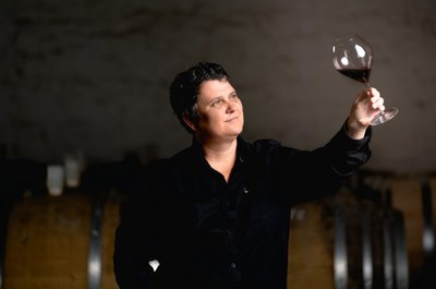 Eine Person in schwarzer Kleidung inspiziert ein Weinglas mit Rotwein, umgeben von Holzfässern im Hintergrund.