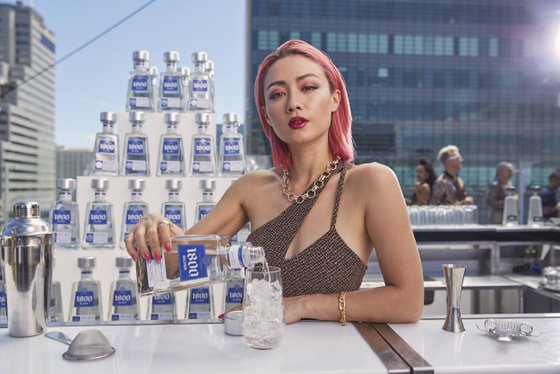 Eine Frau mit pinken Haaren gießt Tequila in ein Glas mit Eis, während eine Flasche 1800 Silver im Vordergrund steht.