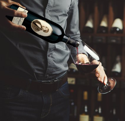 Ein Mann in einem grauen Hemd gießt roten Wein aus einer Flasche in ein Glas, während er eine entspannte Haltung einnimmt.