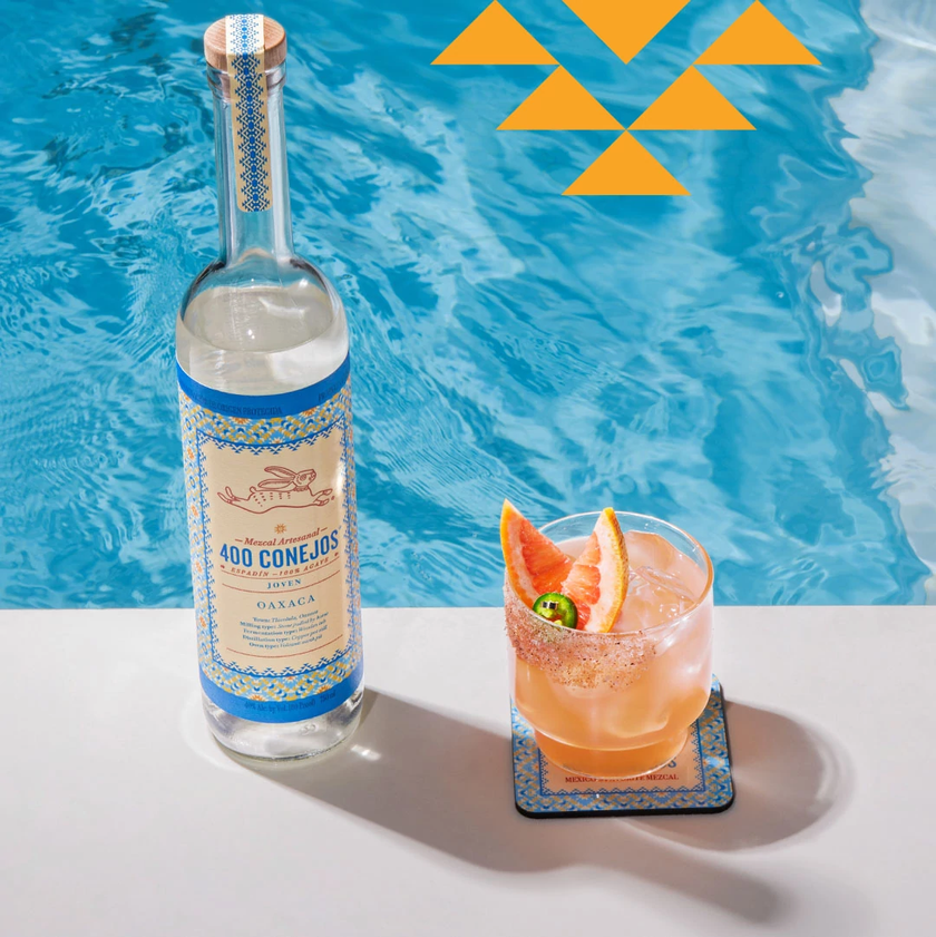 Eine Flasche Mezcal 400 Conejos steht neben einem Cocktailglas mit einer fruchtigen Garnitur am Rand eines blauen Pools.