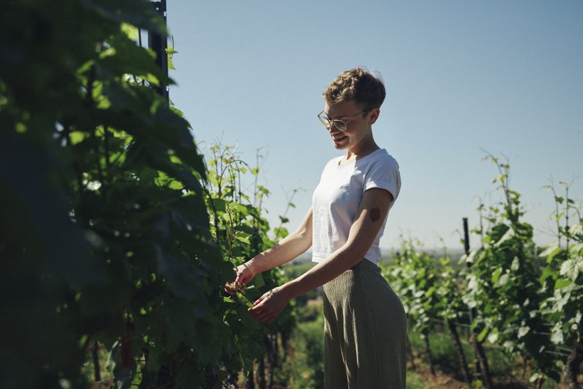 Eine lächelnde Frau in einem Weinberg, die sanft an den Reben arbeitet, umgeben von üppigem Grün und klarem Himmel.