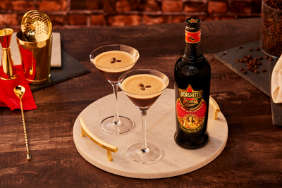 Zwei Martini-Gläser mit schaumigem, braunem Espresso-Martini, garniert mit Kaffeebohnen, stehen auf einem runden, grauen Tablett. Daneben eine Flasche Borghetti-Likör. Im Hintergrund sorgt warmes Bar-Ambiente mit goldfarbenem Bar-Zubehör für eine stilvolle Atmosphäre.