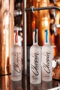 Drei Flaschen Chopin Wodka stehen auf einem glänzenden Tisch, umgeben von einer warmen, kupferfarbenen Brennerei.