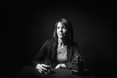 Eine Frau mit schulterlangen Haaren sitzt an einem Tisch und hält ein Glas Whisky in der Hand, während sie nachdenklich schaut.