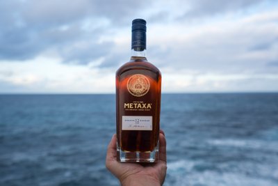 Eine Hand hält eine Flasche Metaxa 12 Sterne vor einem weiten, blauen Meer und einem bewölkten Himmel.
