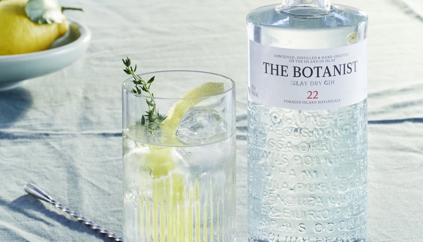 Ein erfrischender Gin-Cocktail mit Zitrone und Kräutern in einem Glas, das auf einem eleganten Tisch neben einer Flasche Botanist steht.