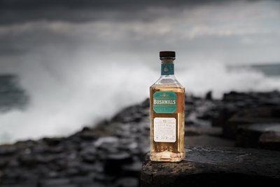 Die Flasche Bushmills Whiskey leuchtet golden im Kontrast zu den grauen Wellen und dem dramatischen Himmel im Hintergrund.