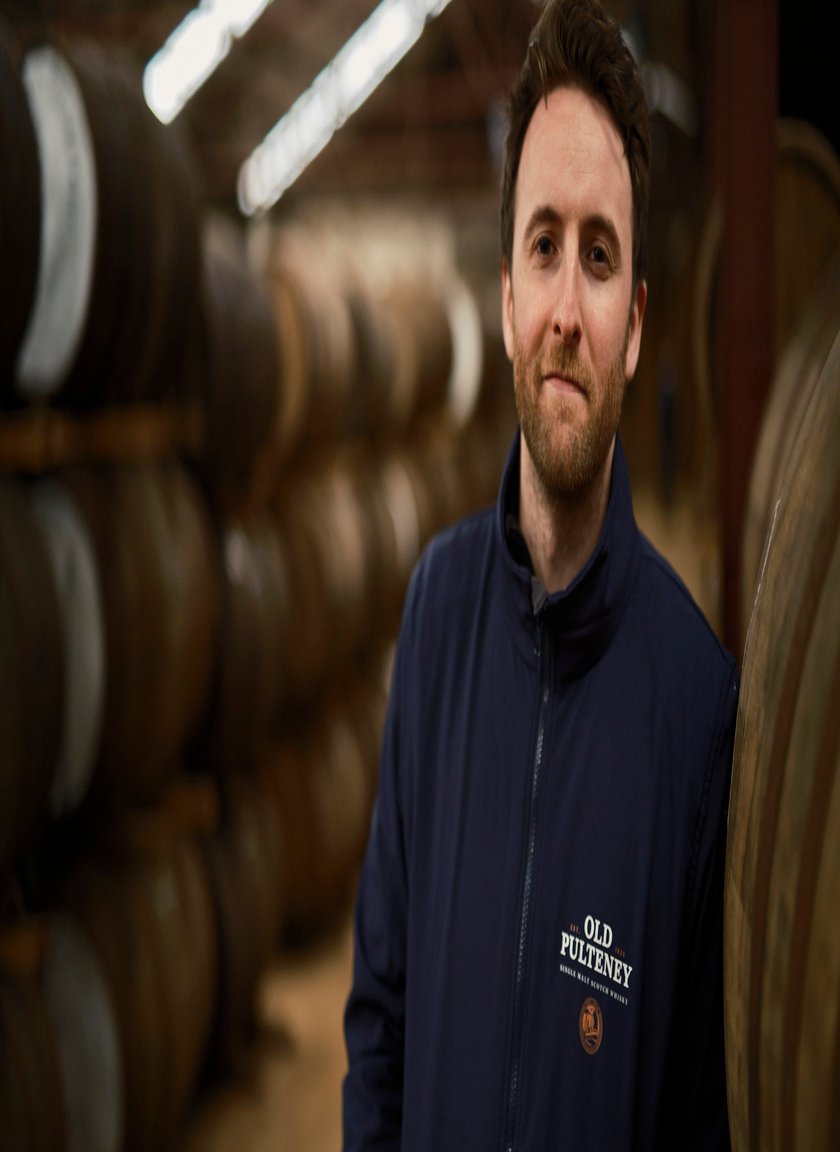 Ein Mann mit Bart steht in einer Lagerhalle, umgeben von großen Holzfässern, und lächelt leicht in die Kamera.