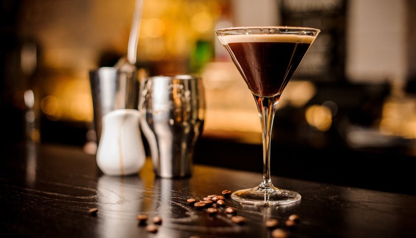 Ein eleganter Cocktail in einem Martiniglas steht auf einem dunklen Tisch, umgeben von Kaffeebohnen und Barutensilien.