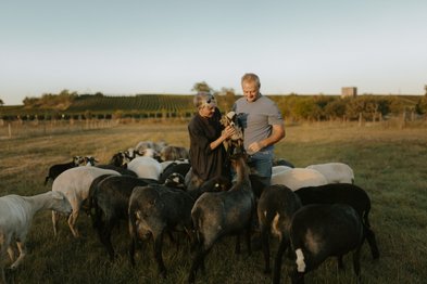 Ein Paar steht in einem Feld voller Schafe und hält ein kleines Zicklein, während sie sich freundlich anlächeln.