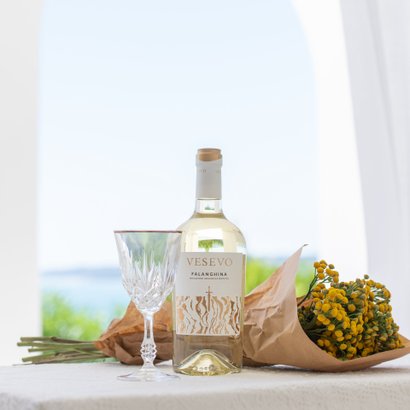 Ein eleganter Tisch mit einer Flasche Vesevo Falanghina, einem Kristallglas und einem Strauß gelber Blumen in braunem Papier.