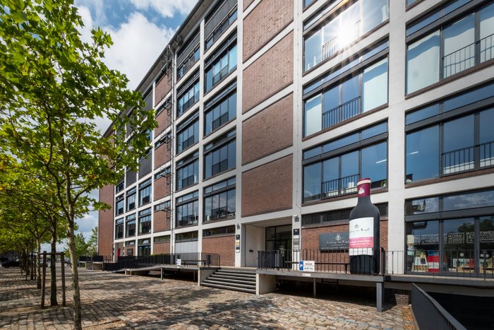 Ein modernes Gebäude mit großen Fenstern und einer auffälligen Weinflasche an der Fassade, umgeben von Bäumen und einem gepflasterten Weg.