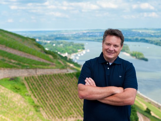 Ein Mann mit verschränkten Armen lächelt vor einer malerischen Weinberglandschaft und einem Fluss im Hintergrund.