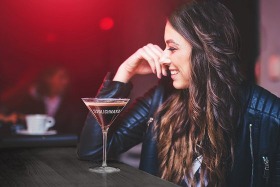Eine lächelnde Frau mit langen, lockigen Haaren sitzt an einem Tisch und genießt einen Cocktail in einem eleganten Glas.