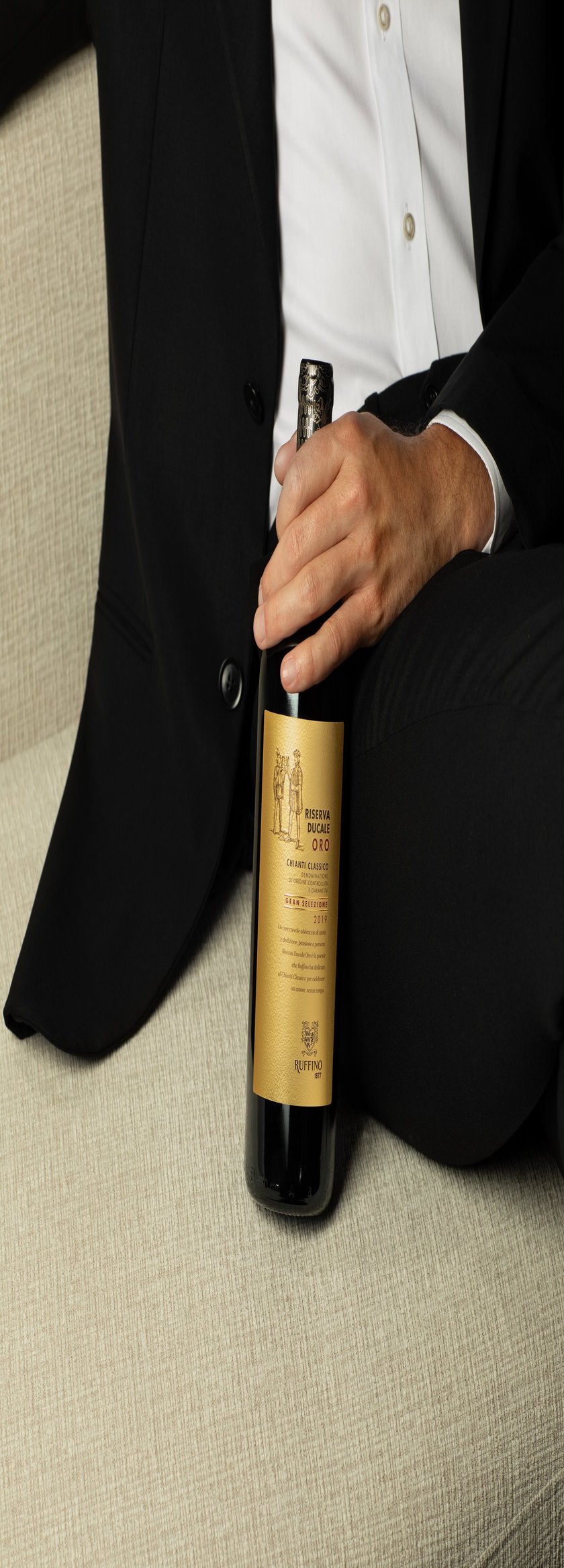 Ein Mann in einem schwarzen Anzug hält eine Flasche Chianti Classico mit goldener Etikette auf einem hellen Sofa.