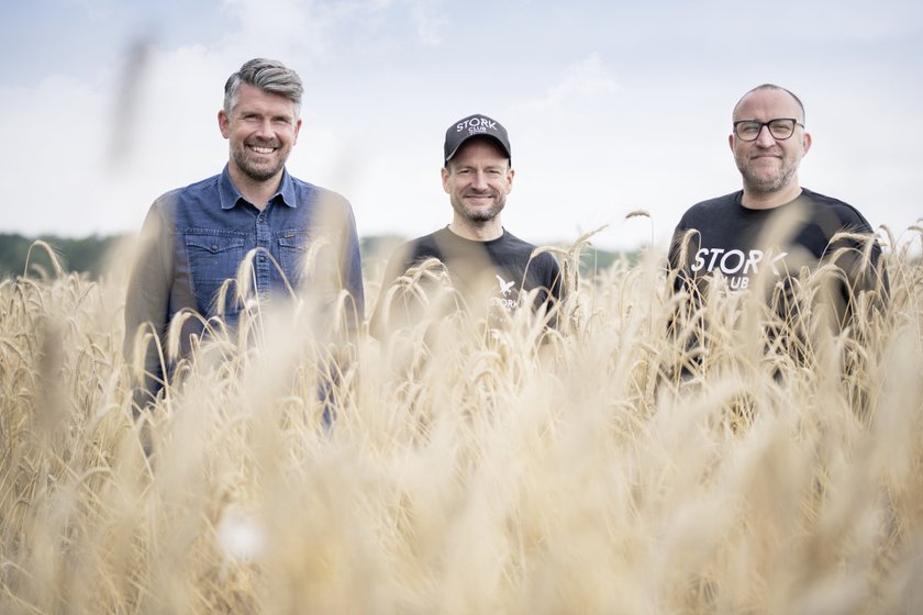 Drei Männer stehen lächelnd in einem goldenen Getreidefeld, umgeben von hohen Halmen unter einem bewölkten Himmel.