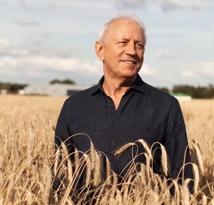 Ein älterer Mann mit grauen Haaren steht in einem goldenen Weizenfeld und lächelt, während er den Himmel betrachtet.
