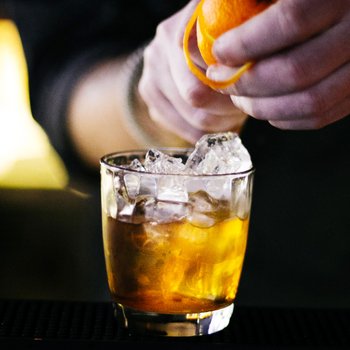 Ein Barkeeper drückt eine Orange über einem Glas mit Eis und einem goldenen Getränk aus, während die Bar im Hintergrund verschwommen ist.
