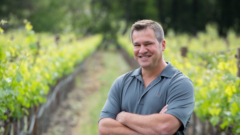 Ein lächelnder Mann steht in einem Weinberg, umgeben von grünen Reben, mit verschränkten Armen und einem freundlichen Ausdruck.