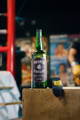 Eine Flasche Proper Twelve Irish Whiskey steht auf einem Holztisch, daneben liegt ein schwarzes Boxband mit gelbem Etikett.