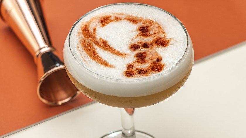 Ein kunstvoll zubereiteter Cocktail in einem Glas, umgeben von einem Kupfermaß und einem Sieb, auf einem modernen Tisch.